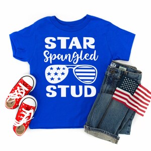 May include: A blue t-shirt with the text "STAR Spangled STUD" in white. The shirt features a pair of sunglasses with stars and stripes. The shirt is paired with blue jeans and a small American flag.