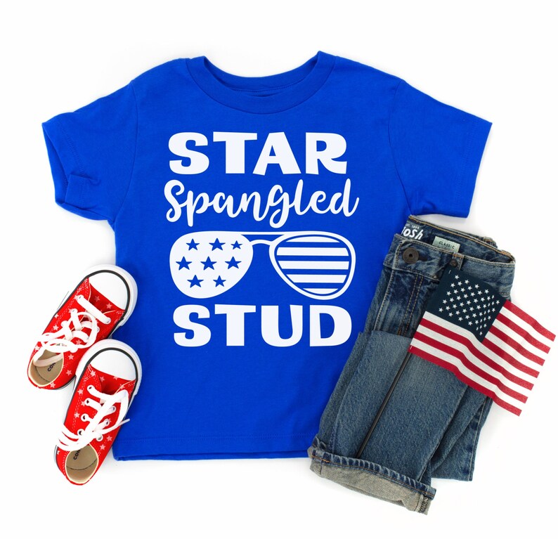 May include: A blue t-shirt with the text "STAR Spangled STUD" in white. The shirt features a pair of sunglasses with stars and stripes. The shirt is paired with blue jeans and a small American flag.