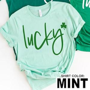 May include: Mint green t-shirt with the word "lucky" in a dark green script font and a green shamrock. The shirt has rolled-up sleeves and is made of a soft material. The text "SHIRT COLOR: MINT" is at the bottom.
