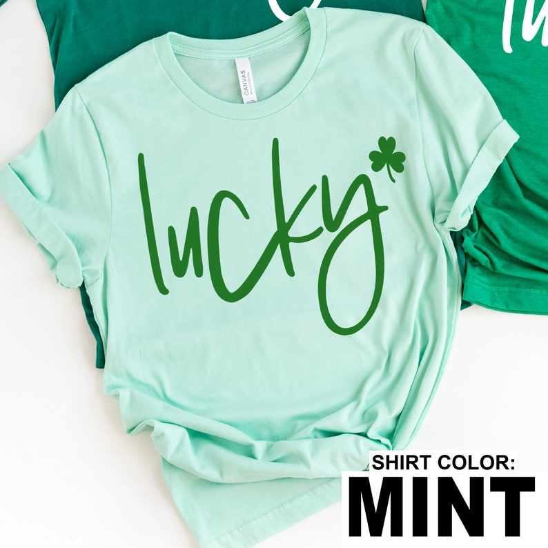 May include: Mint green t-shirt with the word "lucky" in a dark green script font and a green shamrock. The shirt has rolled-up sleeves and is made of a soft material. The text "SHIRT COLOR: MINT" is at the bottom.