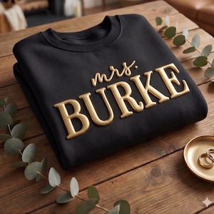 May include: A black crewneck sweatshirt featuring the text "mrs. BURKE" in raised, metallic gold lettering. The garment is folded and displayed on a wooden surface alongside a small gold dish containing two simple gold rings and a sprig of greenery.