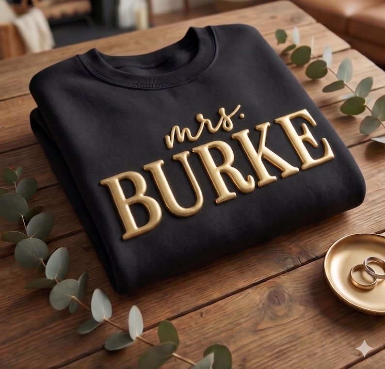 May include: A black crewneck sweatshirt featuring the text "mrs. BURKE" in raised, metallic gold lettering. The garment is folded and displayed on a wooden surface alongside a small gold dish containing two simple gold rings and a sprig of greenery.