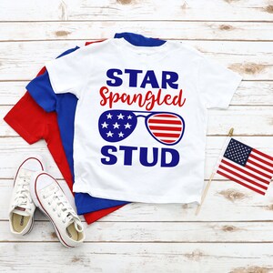 May include: White t-shirt with a red, white, and blue design. The design features the text "STAR Spangled STUD" with sunglasses in the shape of the American flag.