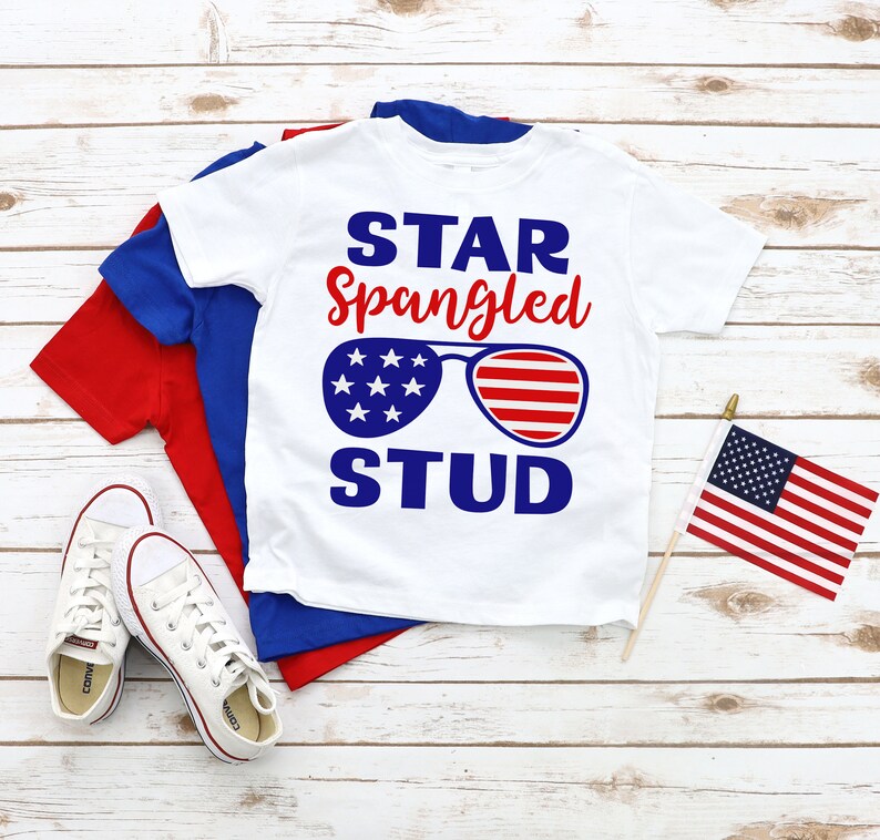 May include: White t-shirt with a red, white, and blue design. The design features the text "STAR Spangled STUD" with sunglasses in the shape of the American flag.