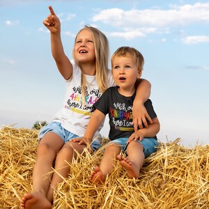 May include: Two children, a girl and a boy, are sitting on a pile of hay. The girl is wearing a white shirt with the text "Watch out 4th grade here I come" and the boy is wearing a black shirt with the text "Watch out Kindergarten here I come". Both shirts have rainbow coloured letters.
