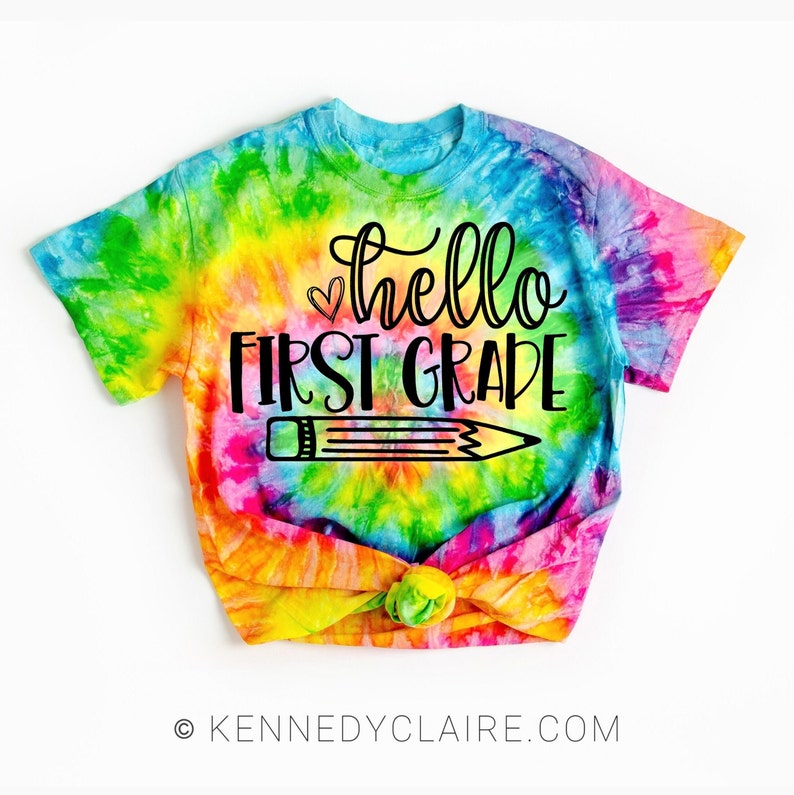 May include: A tie-dye t-shirt with the text "hello first grade" and a pencil graphic. The shirt is a bright rainbow color.