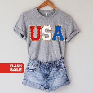 May include: A gray t-shirt with the word "USA" in red, white, and blue sequined letters. The shirt is paired with denim shorts. A red banner with the words "FLASH SALE" is in the lower left corner.
