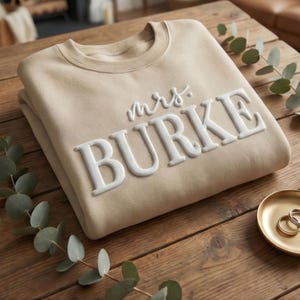 May include: A beige crewneck sweatshirt featuring the white raised text "mrs. BURKE" across the chest. The garment has long sleeves and a ribbed neckline, showcasing a clean design with prominent lettering that stands out against the fabric.