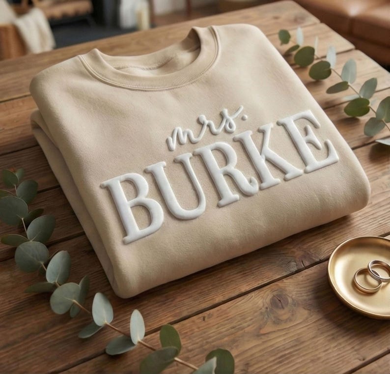 May include: A beige crewneck sweatshirt featuring the white raised text "mrs. BURKE" across the chest. The garment has long sleeves and a ribbed neckline, showcasing a clean design with prominent lettering that stands out against the fabric.