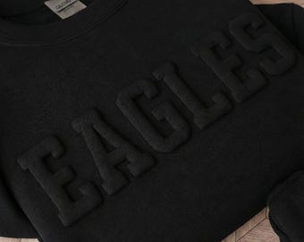 May include: A black crewneck sweatshirt featuring the word EAGLES in large, raised, tonal puff lettering across the chest. The garment has a ribbed neckline and a white interior brand label with the text GLOAN visible near the collar area.