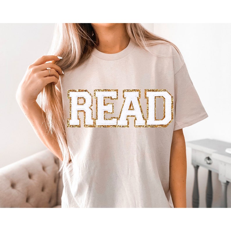 May include: A beige t-shirt with the word "READ" in white letters outlined in gold glitter.