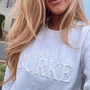 May include: A white long-sleeve crewneck sweatshirt featuring the word "mrs." in a cursive font above the name "BURKE" in large, raised, block-style lettering. The text is monochromatic, matching the white fabric of the garment.
