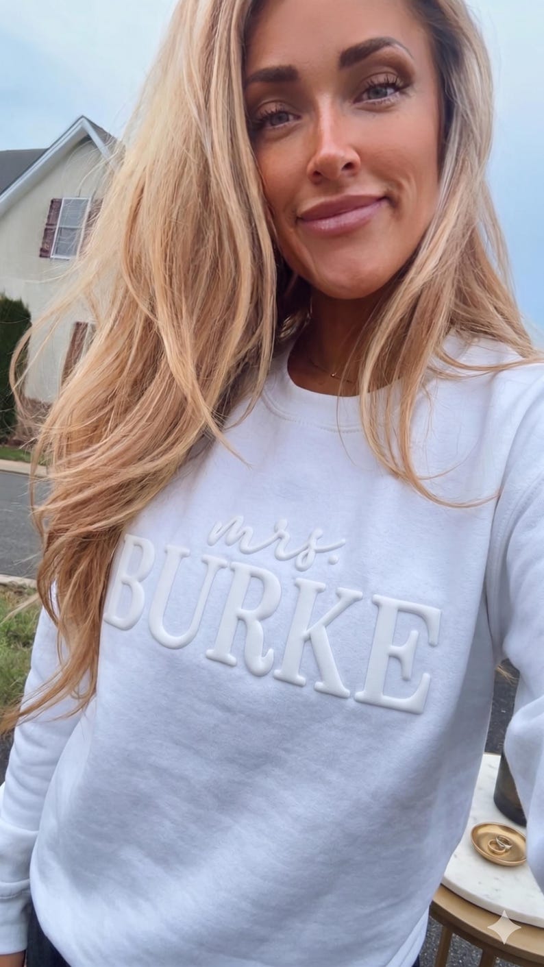 May include: A white long-sleeve crewneck sweatshirt featuring the word "mrs." in a cursive font above the name "BURKE" in large, raised, block-style lettering. The text is monochromatic, matching the white fabric of the garment.