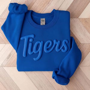May include: A royal blue long-sleeve sweatshirt featuring the word Tigers in a raised, puffy, tonal script font across the chest. The garment has a crew neckline and a Gildan brand label visible at the inner collar, with sleeves folded at the cuffs.