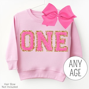 May include: Pink sweatshirt with gold glitter lettering that says "ONE". The sweatshirt is on a white background.  A pink bow is on top of the sweatshirt.