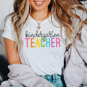 Puede incluir: Camiseta blanca con el texto "kindergarten TEACHER" en colores arco&iacute;ris.