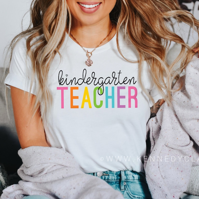 Puede incluir: Camiseta blanca con el texto "kindergarten TEACHER" en colores arco&iacute;ris.