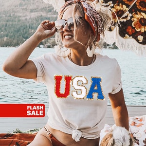 May include: A white t-shirt with a glittery red, white, and blue "USA" graphic. The shirt is knotted at the waist and the model is wearing it with a red, white, and blue bandana and sunglasses.