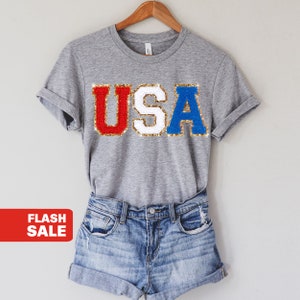 May include: A gray t-shirt with the word "USA" in red, white, and blue glitter letters. The shirt is on a hanger and is paired with denim shorts.