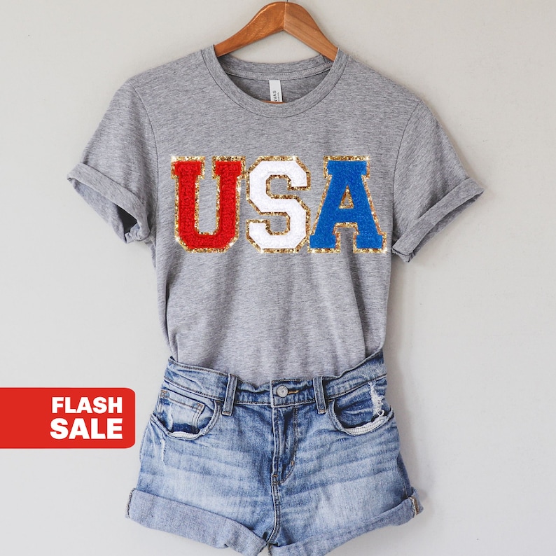 May include: A gray t-shirt with the word "USA" in red, white, and blue glitter letters. The shirt is on a hanger and is paired with denim shorts.