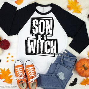 以下が含まれることがあります： 「SON OF A WITCH」の文字が太字で縁取りされた、白と黒のラグランスタイルのシャツ。シャツはブルージーンズとオレンジ色のスニーカーと組み合わされています。カボチャ、葉、クモなど、ハロウィーンをテーマにした装飾が服を囲んでいます。