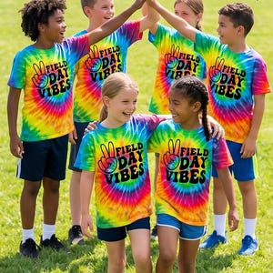Puede incluir: Ni&ntilde;os con camisetas te&ntilde;idas con la t&eacute;cnica tie-dye con el texto "FIELD DAY VIBES" y pantalones cortos azul marino. Las camisetas son un remolino de colores del arco&iacute;ris. El texto "READY FOR THE FIELD!" est&aacute; escrito encima de los ni&ntilde;os.