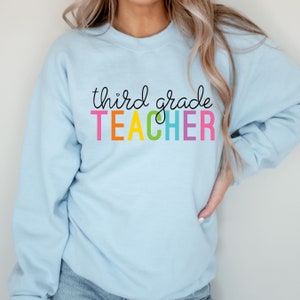 Puede incluir: Una sudadera azul claro con el texto "third grade TEACHER" en colores arco&iacute;ris.