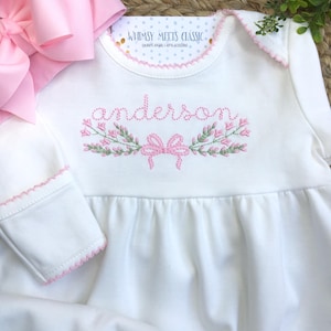 May include: White baby dress with pink embroidered floral wreath and the name "anderson". The dress has pink trim and a matching pink bow. The text "Whimsy Meets Classic" is at the top.