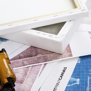 May include: Close-up of a white canvas stretched over a wooden frame. The canvas is stapled to the frame and the staples are visible. The canvas is lying on top of a printed sheet of paper with the text "ARTPRINTCANVAS" visible. A yellow staple gun is in the foreground.