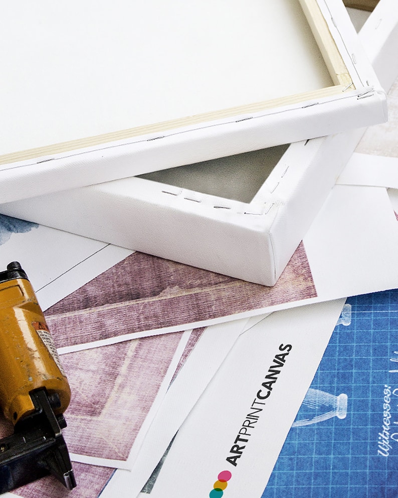 May include: Close-up of a white canvas stretched over a wooden frame. The canvas is stapled to the frame and the staples are visible. The canvas is lying on top of a printed sheet of paper with the text "ARTPRINTCANVAS" visible. A yellow staple gun is in the foreground.