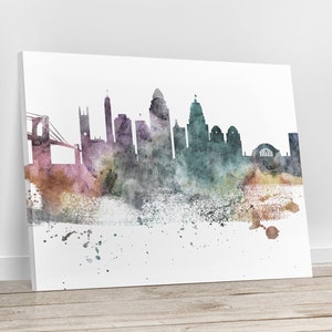May include: A watercolour painting of a city skyline, featuring a bridge and various buildings in shades of purple, green, and brown. The artwork is displayed on a white canvas, leaning against a wall, with a wooden floor in the foreground.