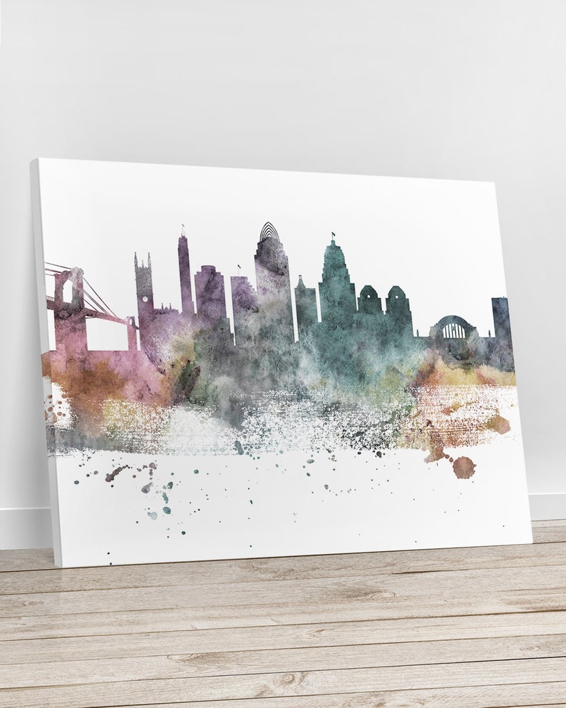 May include: A watercolour painting of a city skyline, featuring a bridge and various buildings in shades of purple, green, and brown. The artwork is displayed on a white canvas, leaning against a wall, with a wooden floor in the foreground.