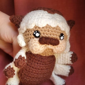 May include: A crocheted stuffed animal resembling a brown and white bison with black eyes. The animal has a fluffy white mane and a brown body.