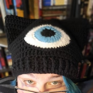 May include: A black crocheted beanie with a large white eye with a blue iris and black pupil. The beanie has cat ears.