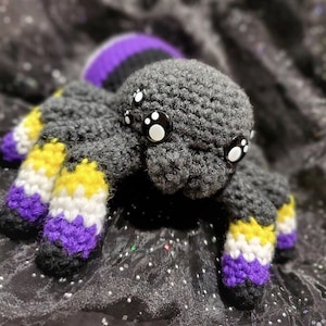 May include: A crocheted spider plush toy with black, purple, yellow, and white stripes on its legs. The spider has large black eyes with white pupils.