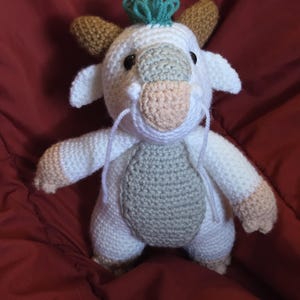 May include: A handmade crochet creature in white, beige, and gray. The plush toy has a round body, small arms and legs, and a detailed face with a light pink nose and teal accents. The toy is sitting on a red surface.