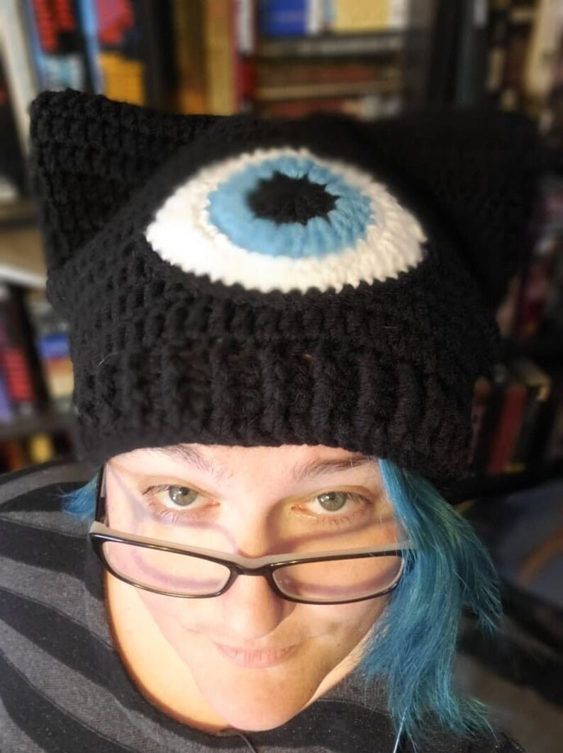 May include: A black crocheted beanie with a large white eye with a blue iris and black pupil. The beanie has cat ears.