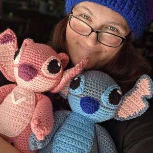 May include: A woman wearing a blue and purple crocheted hat holds two crocheted Stitch plush toys. One Stitch is pink and the other is blue.