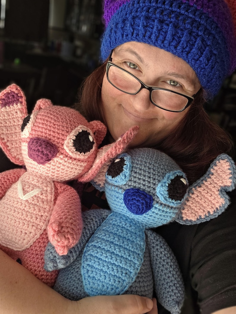 May include: A woman wearing a blue and purple crocheted hat holds two crocheted Stitch plush toys. One Stitch is pink and the other is blue.
