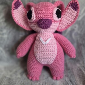May include: Pink crocheted plush toy with large eyes, a purple nose, and white stitching on the chest. The toy is shaped like a cartoon character with large ears.