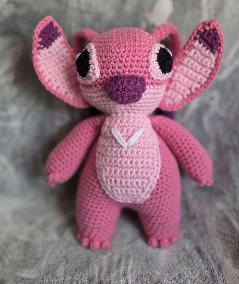 May include: Pink crocheted plush toy with large eyes, a purple nose, and white stitching on the chest. The toy is shaped like a cartoon character with large ears.