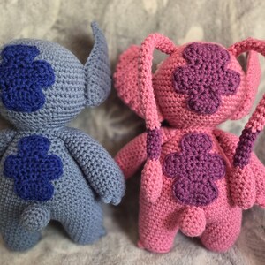 May include: Two crocheted plush toys, one blue and one pink, with embroidered flower designs. The blue toy has a long tail and the pink toy has two long tails.