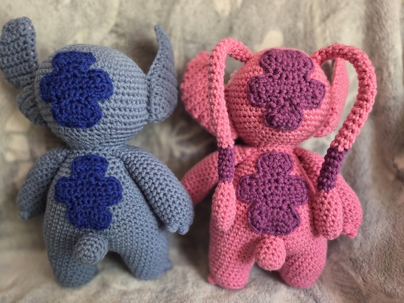 May include: Two crocheted plush toys, one blue and one pink, with embroidered flower designs. The blue toy has a long tail and the pink toy has two long tails.
