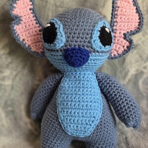 May include: A blue and pink crocheted Stitch plush toy with black eyes and a blue nose.