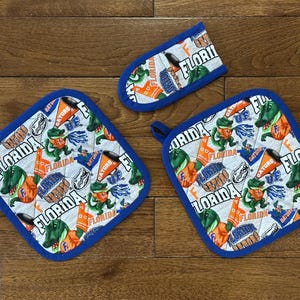 May include: Three blue and white pot holders with a Florida Gators theme. The pot holders feature the team's logo, mascot, and the words "Florida Gators".