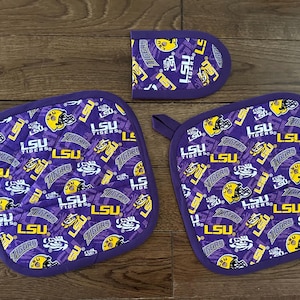 May include: A set of three quilted kitchen items featuring a purple background with yellow and white LSU Tigers logos and helmets. Includes two square pot holders and a small, rounded pot holder. Each item has a purple border and a hanging loop.