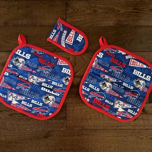 May include: A set of three Buffalo Bills themed kitchen items: two square pot holders and one oven mitt. The items are blue with a red trim and feature the team's logo and name. The pot holders have a loop for hanging.