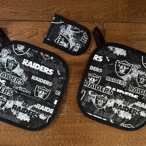 May include: Black and white Raiders-themed kitchen set, including two square pot holders and a small oven mitt. The set features the Raiders logo and team name in a repeating pattern. Each piece has a black loop for hanging.