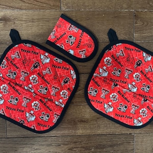 May include: A set of three kitchen textiles featuring a red background with a repeating pattern of Texas Tech logos and text. The set includes two square pot holders and an oven mitt, all with black trim.