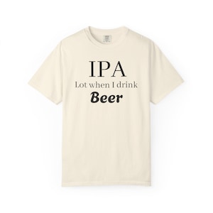 May include: A cream-coloured short-sleeve t-shirt featuring black text printed on the front. The text reads IPA, followed by the phrase Lot when I drink, and the word Beer in a bold, cursive-style font centred below the other lines of text.
